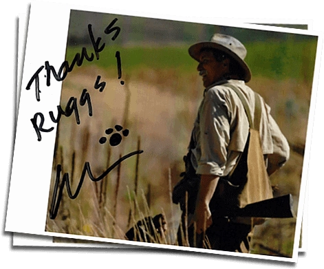 A person in outdoor clothing and hat stands in a grassy field, holding a shotgun. The photo is signed with "Thanks Ruggs!" and a paw print next to a signature.