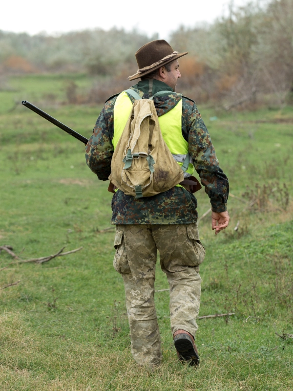 A person wearing camouflage clothing, a yellow vest, and a brown hat walks on grass carrying a backpack and a shotgun.
